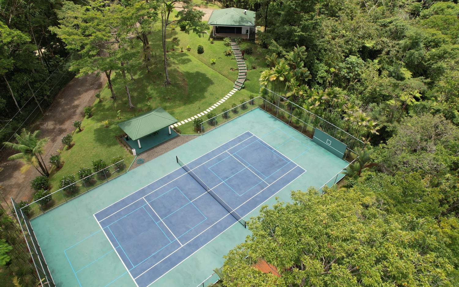 The court at Costa Verde Estates marked for both tennis and pickleball, with a shaded pavilion beside it and the fitness center on the rise above